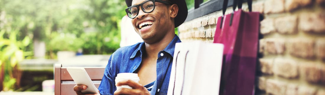 Man sat at a cafe table with shopping bags, a coffee and his phone.